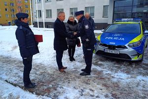 Zdjęcie przedstawia Panią Komendant, policjanta odbierającego dokumenty od Burmistrza Kłobucka, samorządowców oraz oznakowany radiowóz przed budynkiem komendy.