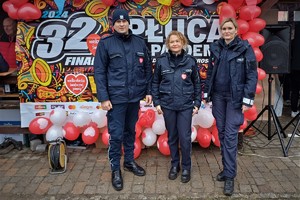 dwie policjantki i policjant w mundurach stoją przy plakacie Wielkiej Orkiestry Świątecznej Pomocy